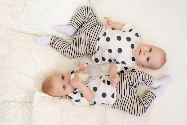 dois gêmeos irmão e irmã 8 meses de idade deitam-se na cama de pijama e bebem leite de uma mamadeira, conceito de comida para bebês, vista superior, conceito de amizade, lugar para texto - Foto, Imagem