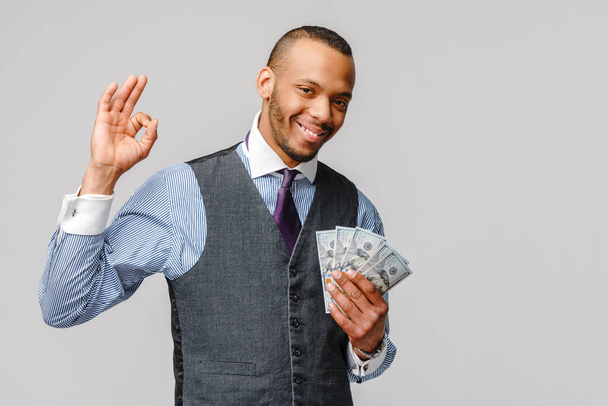 Portret podekscytowanego młodego, afrykańskiego Amerykanina trzymającego gotówkę i pokazującego znak OK na jasnoszarym tle - Zdjęcie, obraz