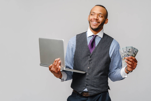 Retrato de animado jovem afro-americano segurando dinheiro em dinheiro e laptop PC sobre fundo cinza claro
 - Foto, Imagem