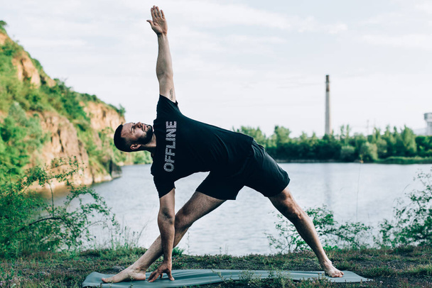 Inspired man doing yoga asanas on quarry lake. Young citizen exercising outside and standing in yoga side angle pose. Fitness outdoors and life balance concept. - Photo, Image