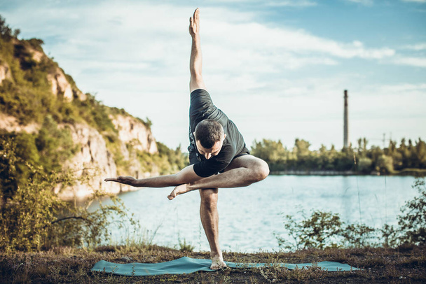 Un beau jeune homme barbu qui fait du yoga sur fond de lac de carrière. Journée internationale de Yoga concept. - Photo, image