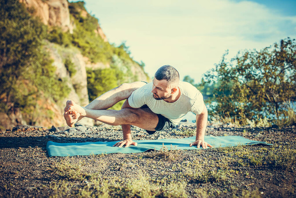 Ajustar al hombre musculoso haciendo yoga junto al agua sobre la hierba con las manos estiradas. Luz solar naranja en el lago de la cantera. Entrenamiento saludable de la columna vertebral. Día Internacional del Yoga
 - Foto, Imagen