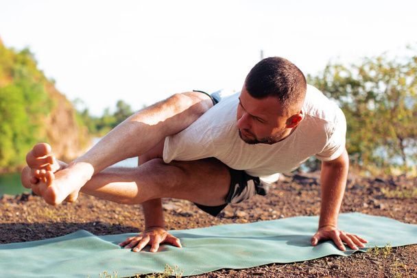 's Ochtends en' s avonds doet een knappe jongeman yoga-oefeningen aan de oever van een steengroeve in de natuur in de zomer buiten. Gezond concept van een actieve levensstijl. - Foto, afbeelding