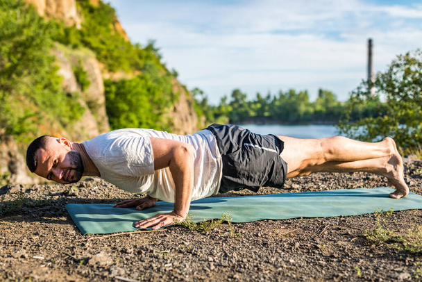 Sabah ve akşam yakışıklı genç bir adam yazın açık havada taş ocağı gölünün kıyısında yoga egzersizi yapıyor. Sağlıklı aktif yaşam tarzı kavramı. - Fotoğraf, Görsel