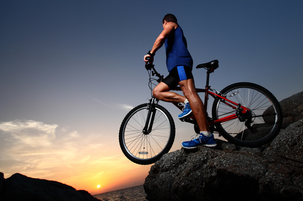 Молодий спортсмен стоїть на скелі з велосипедом
 - Фото, зображення
