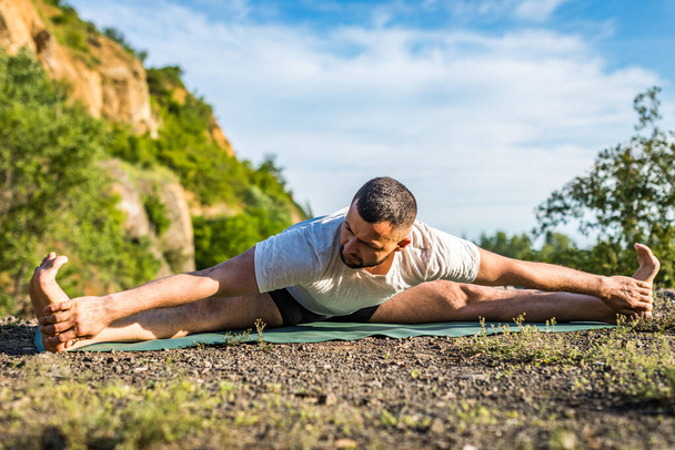 Een jonge fitnessman die buiten yoga doet bij het steengroeve meer. Knappe jongeman met een baard die yoga-oefeningen doet. - Foto, afbeelding