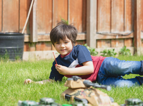 Aktiver Junge legt sich ins Gras und spielt mit Soldaten und Panzerspielzeug im Garten, Kind spielt alleine Krieg und Frieden in heißem, sonnigem Sommer, Fantasie und Entwicklung der Kinder  - Foto, Bild