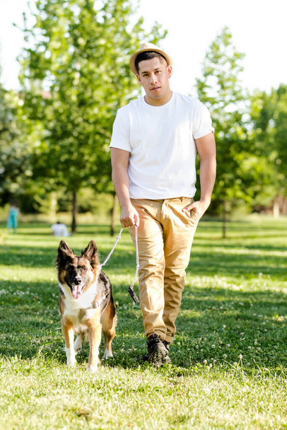 Молодой человек выгуливает собаку в парке. Латинская модель позирует и смотрит на камеру - Фото, изображение