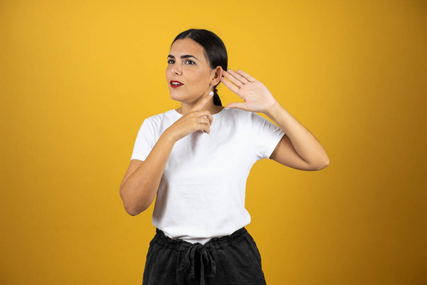 Young pretty woman putting her hand next to her ear pretending she is not listening on yellow background. Wearing white t-shirt, black shorts and red lipstick. - Photo, Image
