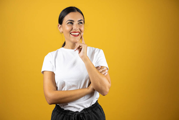 Young woman thinking about something by placing her finger next to her mouth and looking to the right. Standing over an isolated yellow background - Photo, Image