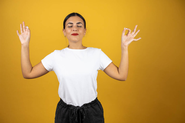 Pretty woman meditating doing the ok sign with both hands. Standing over isolated yellow background - Photo, image