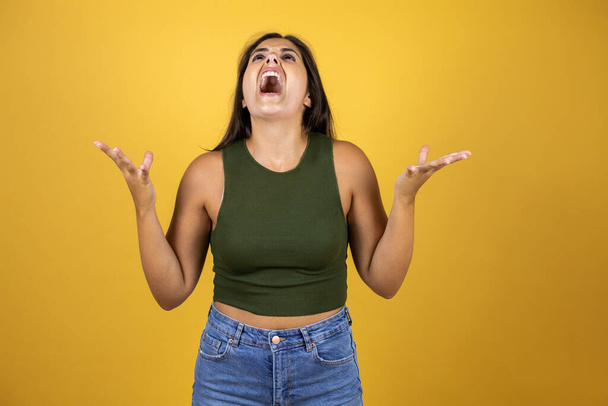 Young beautiful woman standing over yellow isolated background crazy and mad shouting and yelling with aggressive expression and arms raised - Photo, Image