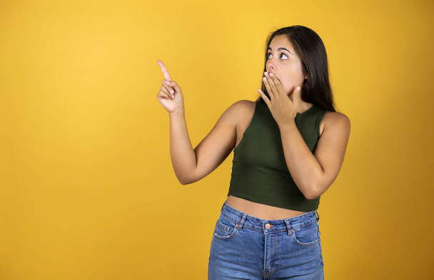 Mladá krásná žena stojící nad žluté izolované pozadí překvapen a její ruka v ústech a ukazuje prstem - Fotografie, Obrázek