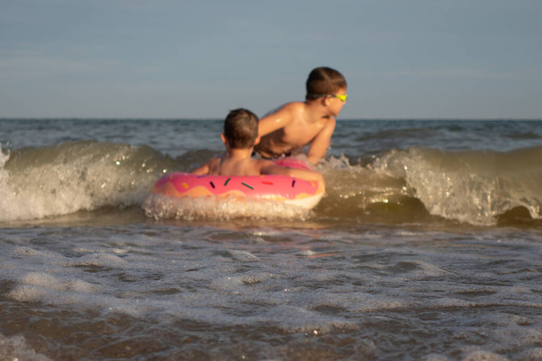 5 ve 10 yaşındaki kardeşler kıyıya yakın denizde yüzüp eğleniyorlar. Donut şeklinde şişme dairesi olan küçük çocuk, yeşil yüzme gözlüklü olan. - Fotoğraf, Görsel