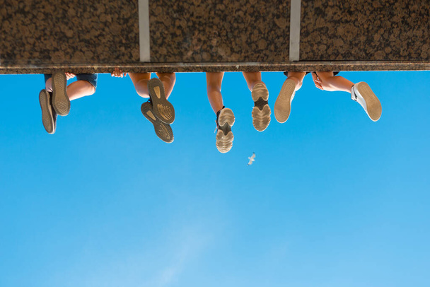 Meninos pernas em sapatos pendurados na ponte contra o céu azul
 - Foto, Imagem
