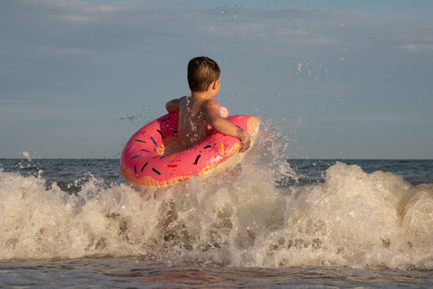 Um menino de 5 anos está nadando e se divertindo no mar perto da costa com um círculo inflável em forma de donut - Foto, Imagem