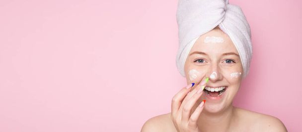 Smiling woman with a towel on her head on a pink background and face cream. Hydration of the skin, skin care face, beauty. - Photo, Image