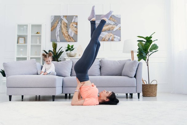 Young mom making stretching exercises and practicing yoga with baby girl at home. Health care and sports concept. - Foto, Imagen