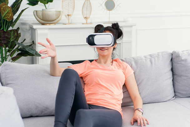 Pretty woman uses augmented reality goggles at home on sofa. Plays games, technologies of the future. - Fotoğraf, Görsel