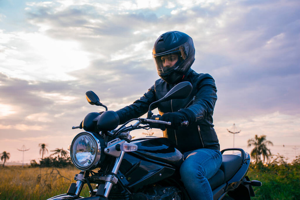 Homem sentado em uma motocicleta preta, vestindo jeans, casaco preto e capacete preto com um fundo do céu com nuvens. - Foto, Imagem