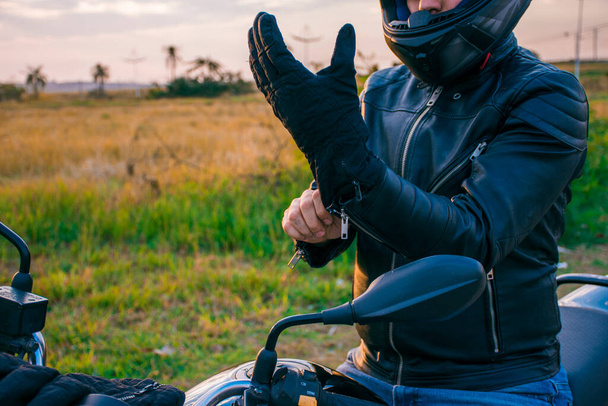 Hombre sentado en una motocicleta negra, vestido con jeans y una chaqueta negra, poniéndose guantes negros con el paisaje en el fondo. - Foto, Imagen