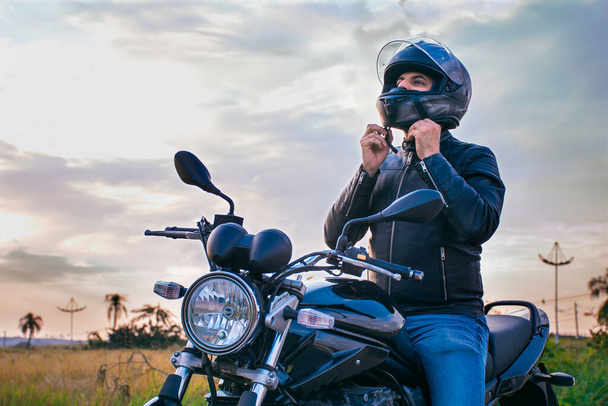 Man zittend op een motorfiets, spijkerbroek en een zwarte jas dragend, zijn helm vastmakend met een landschap op de achtergrond. - Foto, afbeelding