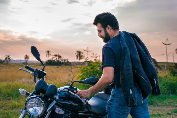 Homme debout à côté d'une moto, vêtu d'un jean et d'une veste noire au-dessus de son épaule, avec un fond naturel. - Photo, image