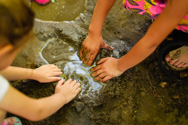 Hands of two little girls making a mud cake out of a muddy puddle as a part of their play with water during the hot summer afternoon - Foto, Imagen