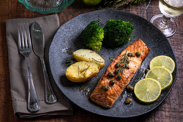 A piece of roasted salmon grilled lemon pepper and salt on a dark plate with broccoli. Wood background - Photo, Image