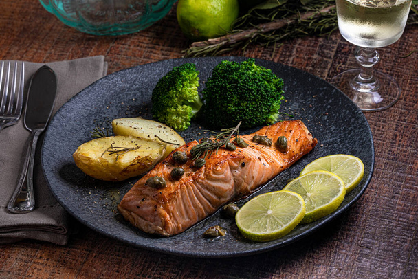A piece of roasted salmon grilled lemon pepper and salt on a dark plate with broccoli. Wood background - Photo, Image