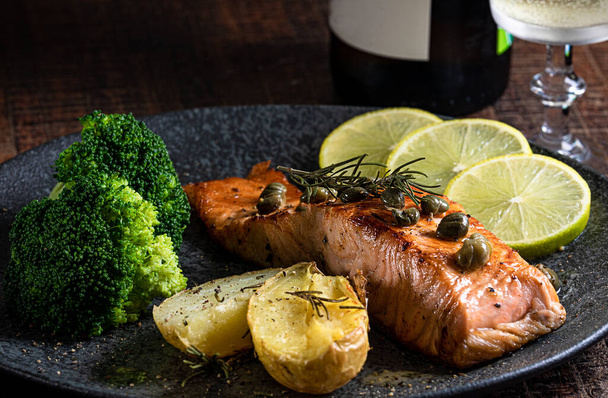 A piece of roasted salmon grilled lemon pepper and salt on a dark plate with broccoli. Wood background - Foto, imagen