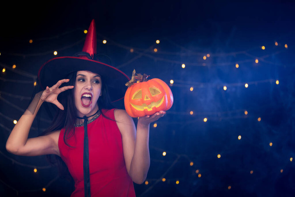 Beautiful girl dressed up in red costume like a witch holding carved pumpkin in Halloween party. - Photo, Image