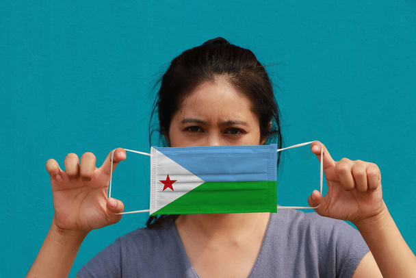 Nainen Djiboutin lippu hygieeninen naamio kädessään ja nosti edessä kasvot vaaleansininen väri tausta. Pieni hiukkanen tai virus korona tai Covid 19 suojaa. Sairauksien torjunnan käsite. - Valokuva, kuva