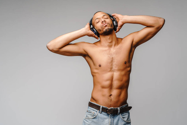 African american young man shirtlesslistening to music on headphones over light grey background - 写真・画像