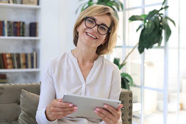 Cheerful middle aged woman sitting on sofa, using computer tablet apps, looking at screen,reading good news in social network, shopping or chatting online - Foto, Bild