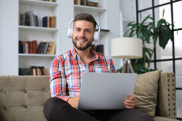Concentrated young freelancer businessman sitting on sofa with laptop, working remotely online at home - Foto, imagen