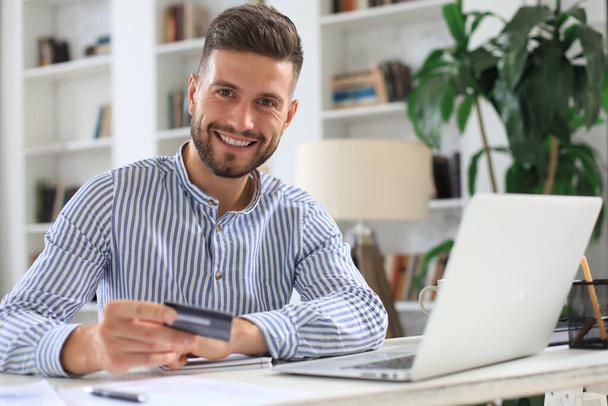 Smiling man sitting in office and pays by credit card with his laptop - Photo, Image