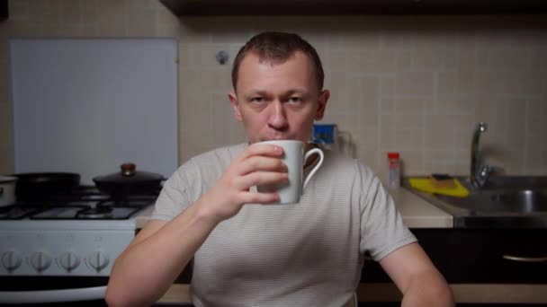 Hombre sentado en casa en la mesa de la cocina y beber café de una taza blanca, movimiento de la cámara - Imágenes, Vídeo