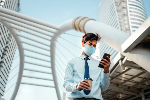 Nuori aasialainen liikemies yllään kirurginen naamio ja käyttämällä älypuhelinta kaupungissa. Terveydenhuolto uudessa normaalissa elämäntavassa käsite - Valokuva, kuva