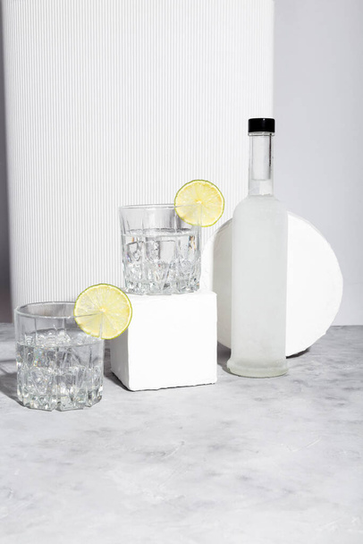 Vodka with ice and lime in two glasses on white stand and bottle of vodka. Shadows and sunlight trends - 写真・画像