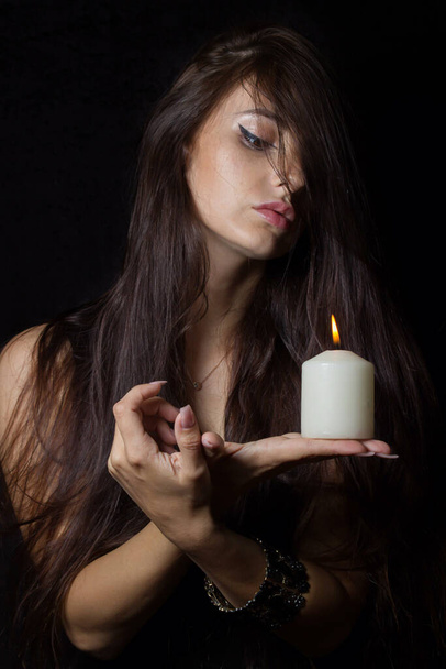 Dziewczyna z płonącymi świecami. Brunetka na czarnym tle trzyma w rękach białą świecę. Mistycyzm i wróżenie. czarna magia - Zdjęcie, obraz