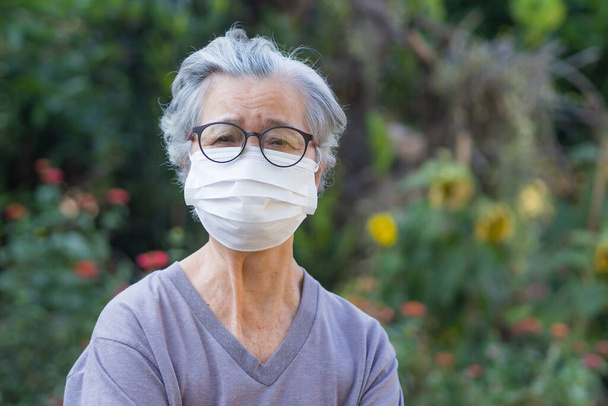 A portrait of an elderly woman wearing a mask while standing in a garden. Mask for protect virus, bacteria, pollen grains. Concept of old people and health care - Photo, image