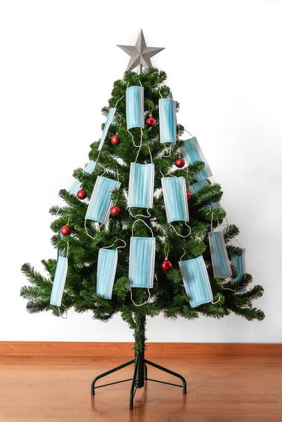 Imagen en color vertical con vista frontal de un árbol de Navidad decorado con bolas rojas, máscaras de coronavirus azul y una gran estrella. Covid y feliz año nuevo concepto. - Foto, imagen