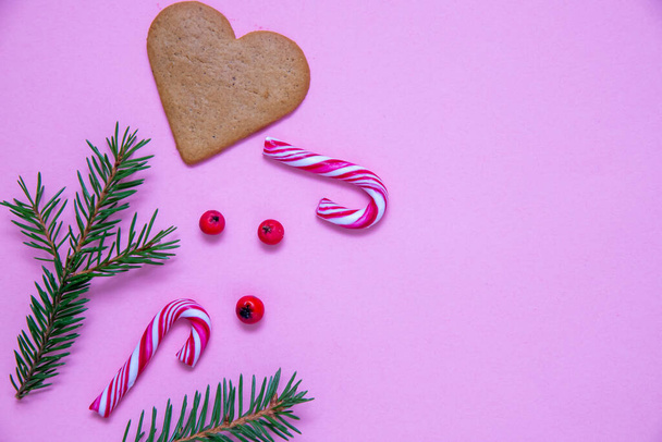 Un plan plat de cannes à bonbons, de branches de sapin, de baies de Noël et d'un biscuit au cœur sur une surface rose - Photo, image
