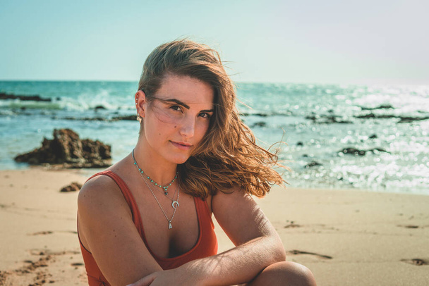 A beautiful European female with an orange swimsuit sitting on sandy beach - Fotó, kép