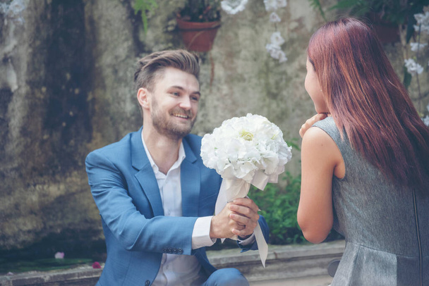 Καυκάσιος όμορφος άντρας δίνει λουλούδια στην Ασιάτισσα κοπέλα του ζητώντας της να τον παντρευτεί στο Γκριν Παρκ. Ζευγάρι εραστής σε ρομαντική ημερομηνία ευτυχισμένη σχέση. Γάμος προτείνει Ημέρα του Αγίου Βαλεντίνου Έννοια. - Φωτογραφία, εικόνα