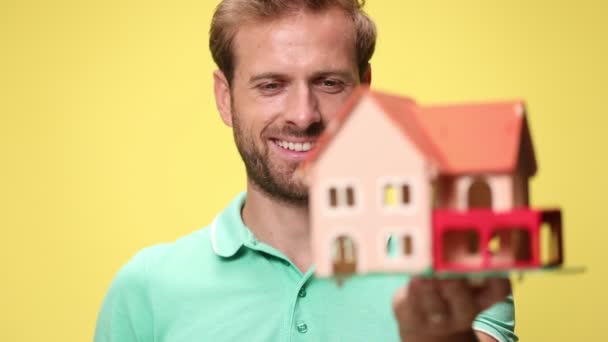 junger Mann bewundert Haus, hält und präsentiert Haus, macht Daumen nach oben Geste und lächelt auf gelbem Hintergrund - Filmmaterial, Video