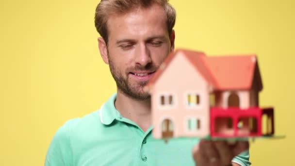 lächelnder junger, lässiger Typ, der das Hausmodell hochhält, empfiehlt und mit dem Finger auf gelbem Hintergrund zeigt - Filmmaterial, Video