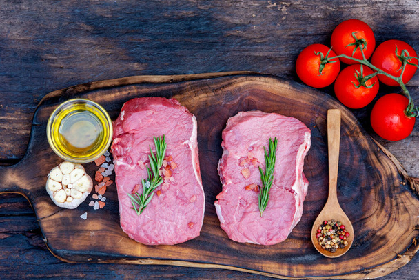 Surowe mięso wołowina stek organiczny świeży składnik na drewnianym tle stołu deski w kuchni z rozmarynu, sól, czosnek, pomidor, czarny pieprz, oliwa z oliwek. Mięso wołowe na drewnianym talerzu do surowego mięsa wołowego - Zdjęcie, obraz