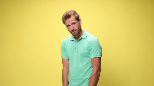 timid young casual man holding hands on hips and standing in a side view pose on yellow background - Footage, Video